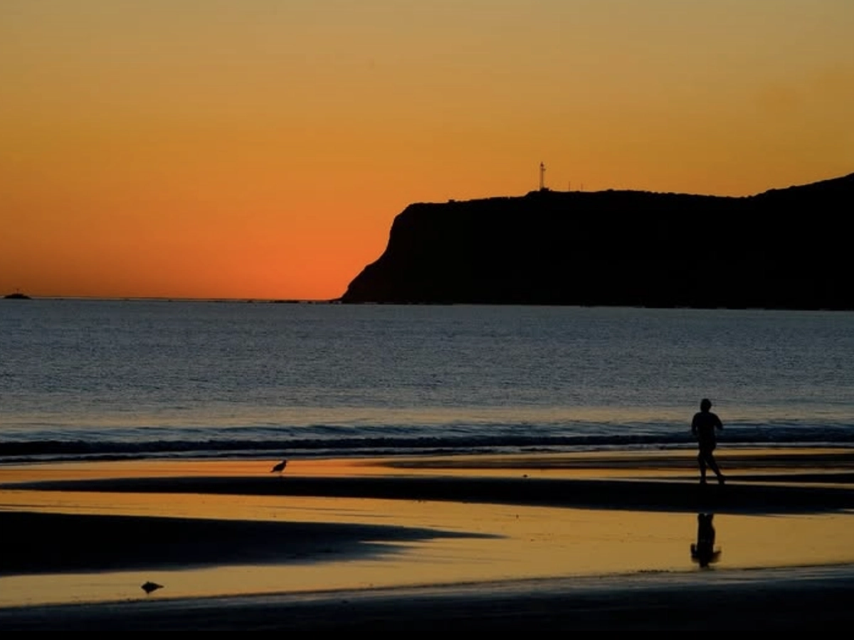 A silhouette of a person walking along the shoreline at sunset, with calm waves reflecting the orange sky and a dark headland in the background.