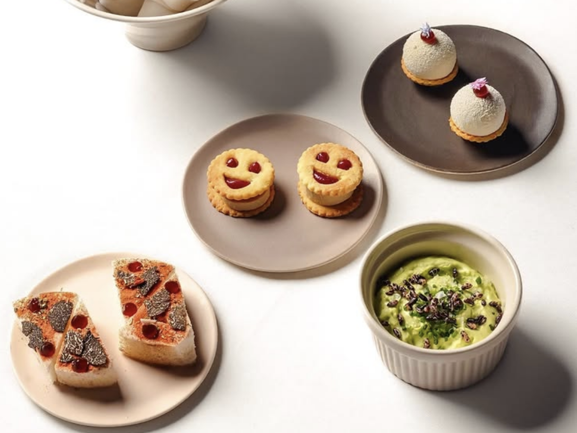 A playful assortment of gourmet bites including truffle toast, smiley face cookies with jam filling, white mousse domes with floral garnish, and a ramekin of creamy green dip topped with herbs and seeds, all artfully plated on neutral-toned dishes.
