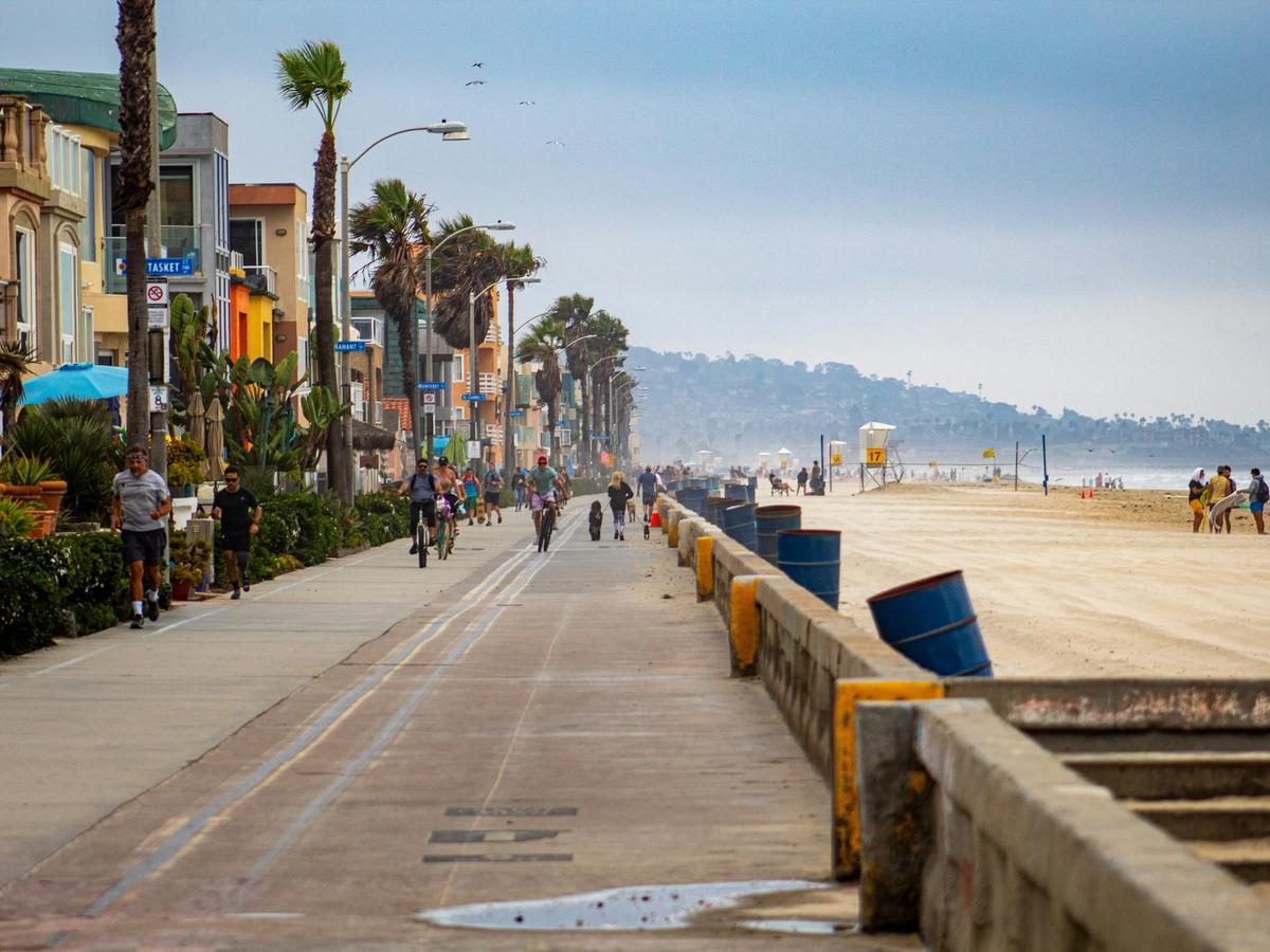 A lively beachside boardwalk with joggers, cyclists, and people walking dogs, lined with colorful houses and palm trees, alongside a wide sandy beach with lifeguard towers in the distance.