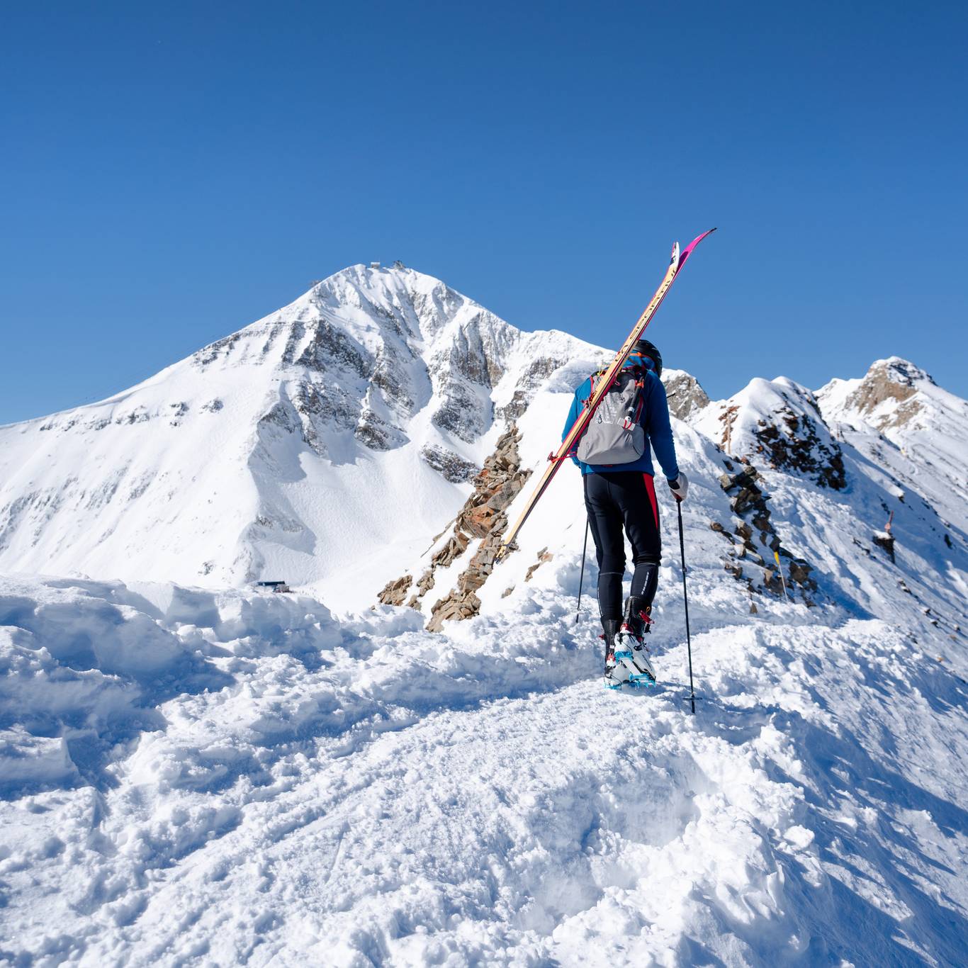 A skier climbs a snowy ridge carrying skis against a clear blue sky. The towering peaks and untouched snow show the challenge and thrill of alpine adventures in Big Sky’s backcountry.
