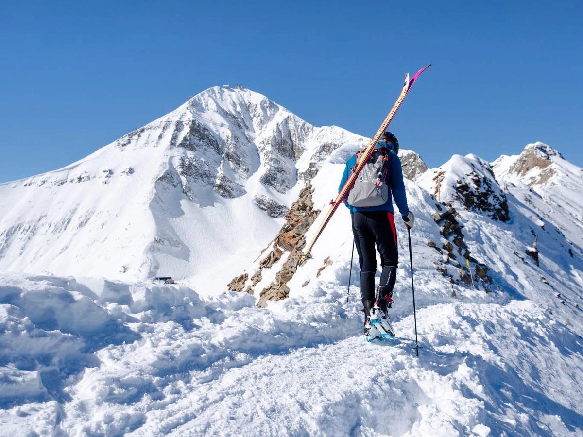 A skier climbs a snowy ridge carrying skis against a clear blue sky. The towering peaks and untouched snow show the challenge and thrill of alpine adventures in Big Sky’s backcountry.