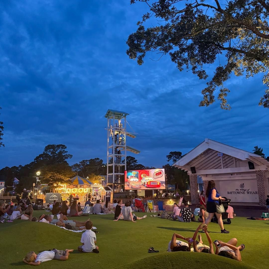 Families relaxing on the lawn at The Village of Baytowne Wharf in Miramar Beach, Florida during an outdoor movie night with shops, attractions, and evening entertainment.