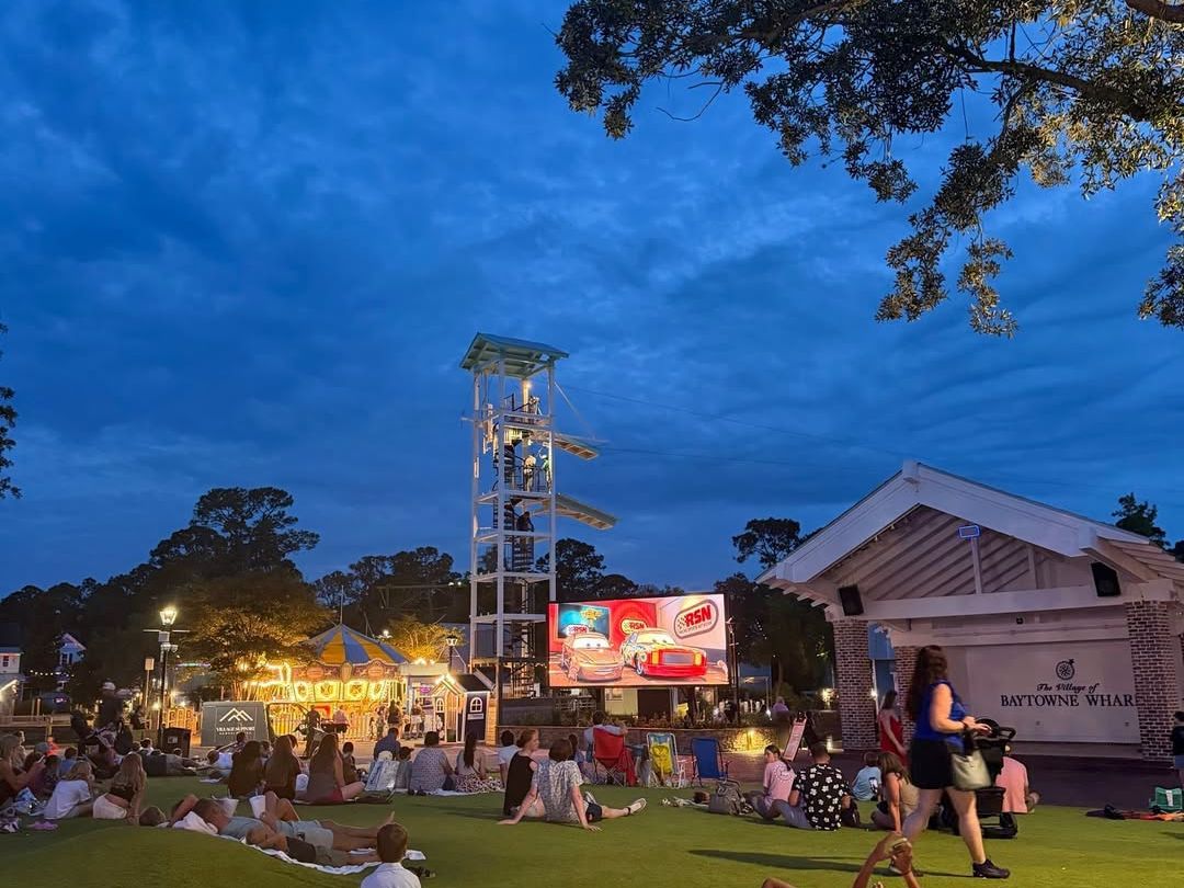 Families relaxing on the lawn at The Village of Baytowne Wharf in Miramar Beach, Florida during an outdoor movie night with shops, attractions, and evening entertainment.