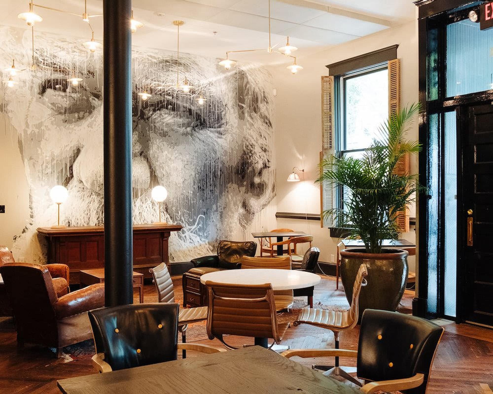 ChatGPT said:  A stylish lounge area features modern leather chairs, round tables, and potted plants, with a striking black-and-white mural of an elderly face covering the back wall.