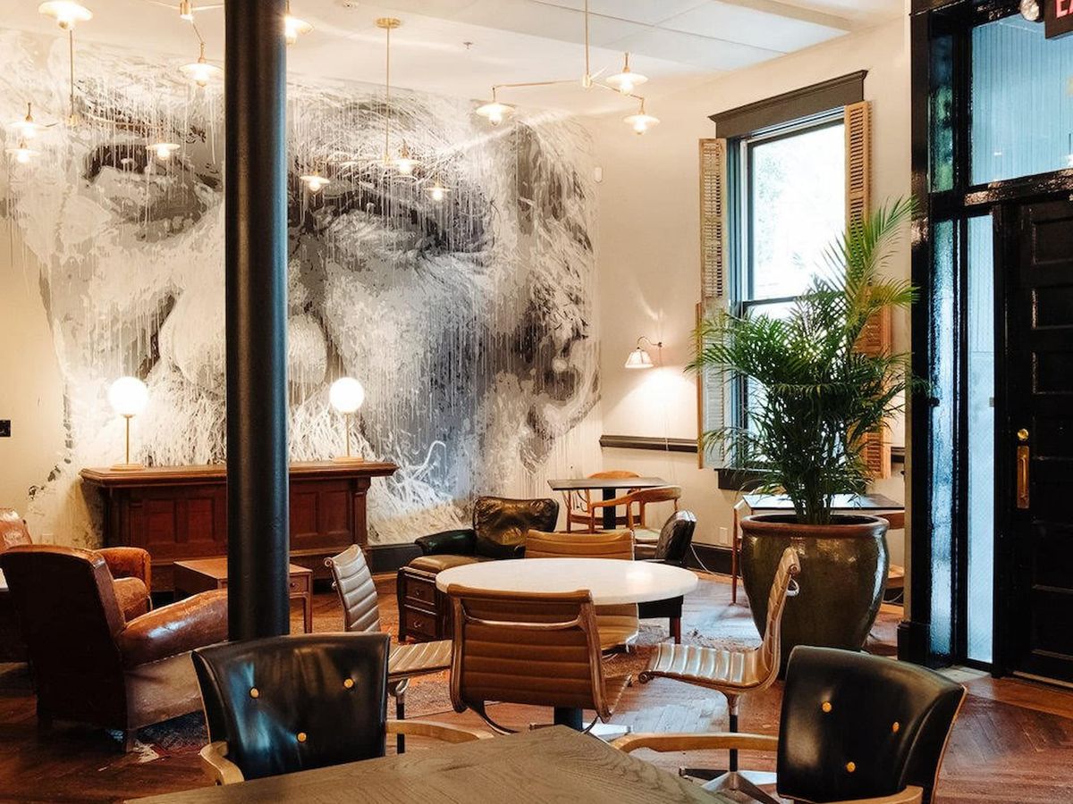 ChatGPT said: A stylish lounge area features modern leather chairs, round tables, and potted plants, with a striking black-and-white mural of an elderly face covering the back wall.