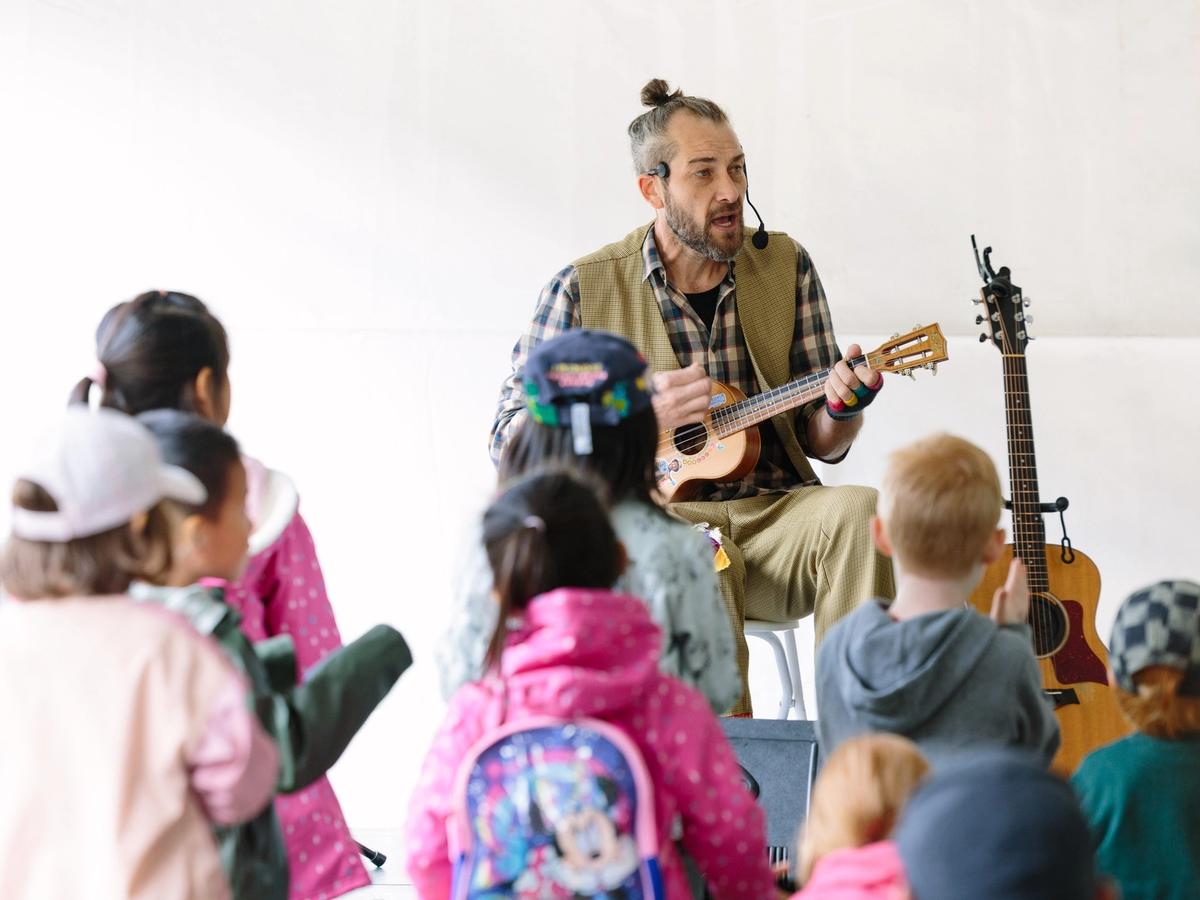 A performer plays music on a ukulele while entertaining a group of children sitting nearby. The kids watch and listen closely during this fun spring festival event. The Whistler Children’s Festival is known for family-friendly performances and activities.
