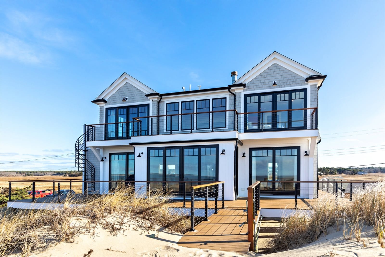 A brand-new Cape Cod vacation rental sits right on the dunes, with a wood walkway cutting through beach grass leading straight up to the front door. The two-story shingle-style home has large black-framed windows on every floor, a spiral staircase on the side, and wide upper and lower decks with cable railings that look out over open marshland and blue sky.