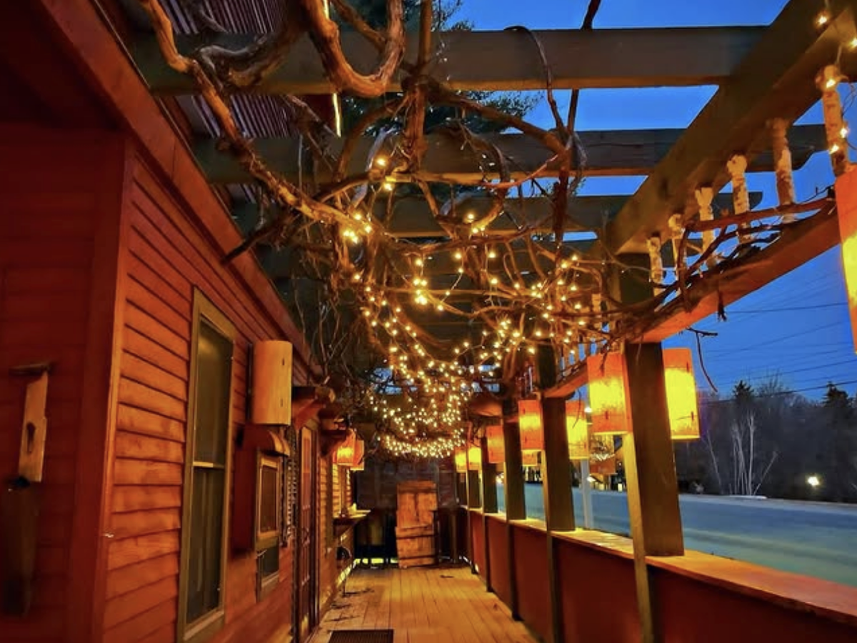 A cozy wooden porch adorned with string lights and hanging lanterns glows warmly at dusk, creating a welcoming ambiance beside a quiet road.