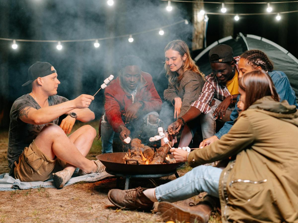 A group of friends gathers around a glowing bonfire, roasting marshmallows under string lights in a cozy campsite setting. Their laughter fills the night as they relax near a tent, surrounded by nature and warm conversation.