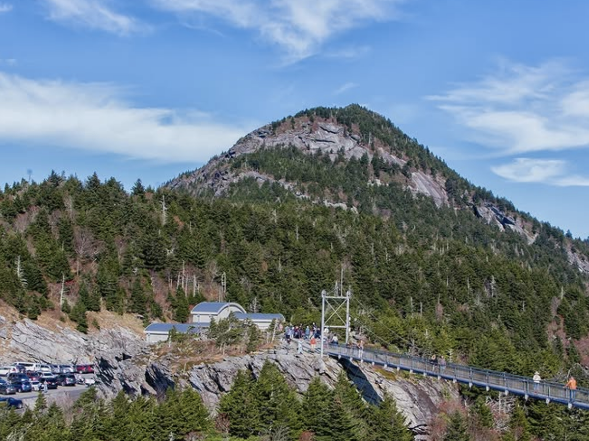 A suspension bridge stretches across a rocky gap at Grandfather Mountain, with a forested mountain peak rising in the background under a blue sky.