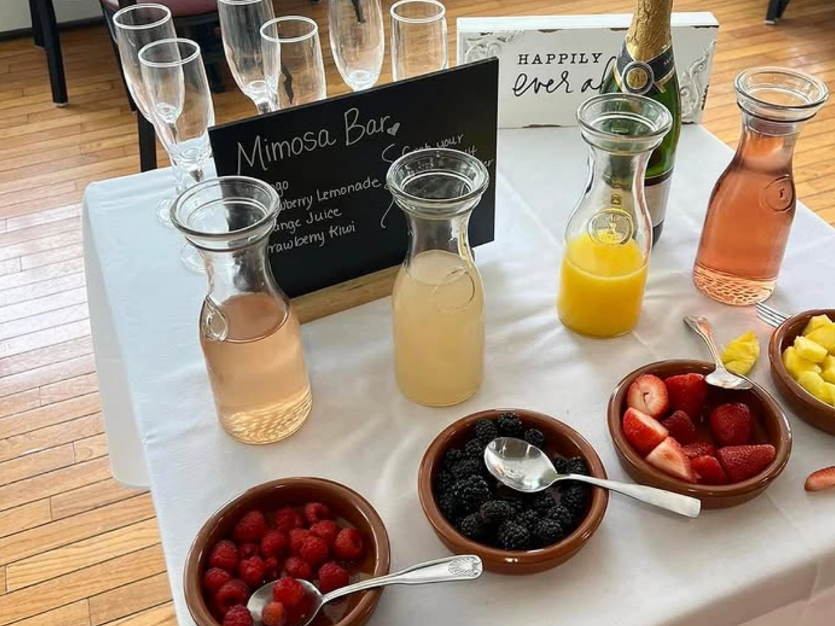 A mimosa bar setup with champagne, carafes of juices, bowls of fresh berries and fruit, and champagne flutes on a white tablecloth.