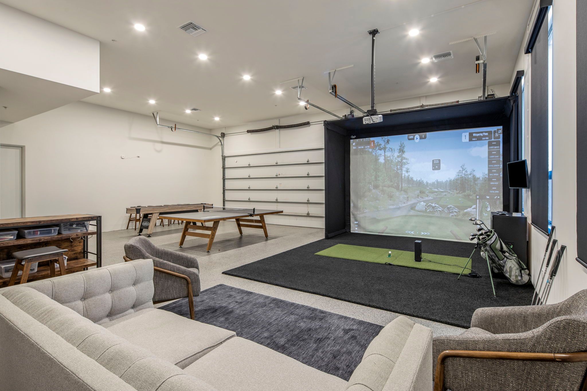 Modern garage-turned-recreation room with a golf simulator, putting mat, ping pong table, and cozy lounge seating under bright recessed lighting.