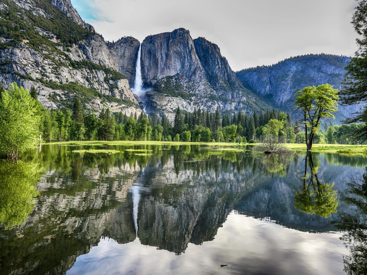 majestic waterfall cascades down granite cliffs into a serene valley surrounded by lush trees, with the entire scene beautifully reflected in the still water below.