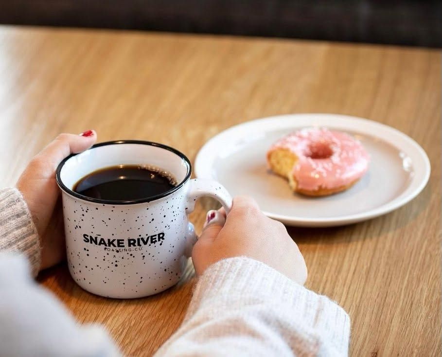 wo hands wrap around a speckled Snake River Roasting Co. mug filled with black coffee on a light wooden table. A pink-frosted doughnut with a bite taken out rests on a white plate nearby, capturing a cozy morning café moment.