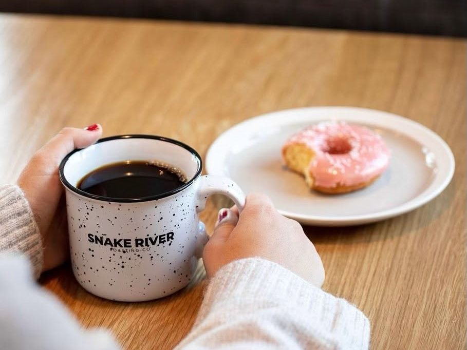 wo hands wrap around a speckled Snake River Roasting Co. mug filled with black coffee on a light wooden table. A pink-frosted doughnut with a bite taken out rests on a white plate nearby, capturing a cozy morning café moment.