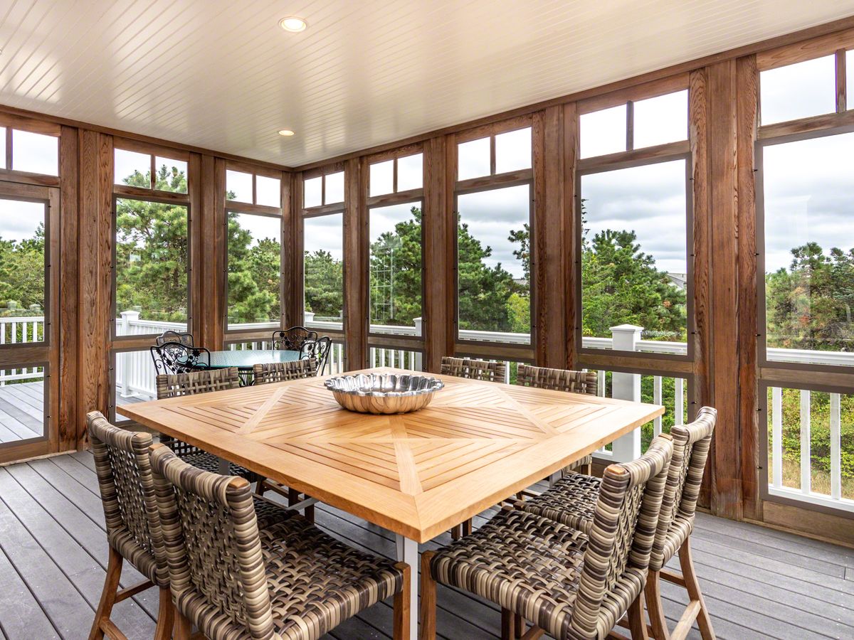 A cozy screened-in porch with wood framing and modern woven chairs surrounds a square dining table, offering a serene view of lush greenery and pine trees beyond the white railing.