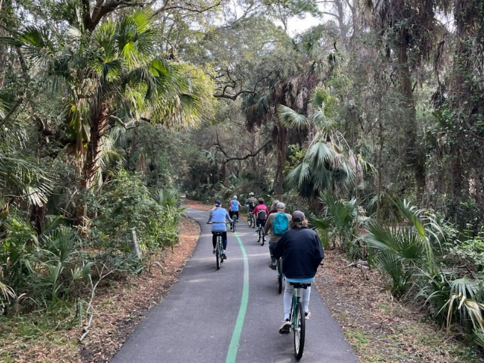 A group of cyclists rides along a shaded forest trail lined with tall palms and thick greenery. The quiet path curves gently ahead, creating a sense of adventure and calm. It’s a relaxed way to explore the island’s natural beauty.