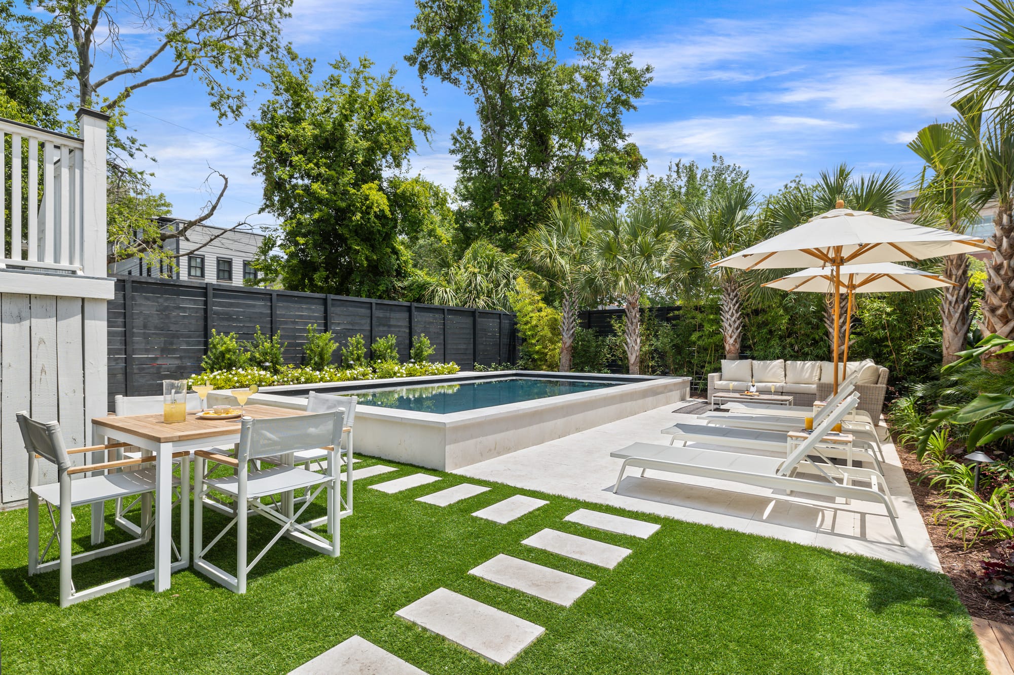 A sunny backyard with a private pool sits surrounded by palm trees and bright green landscaping. The lounge chairs, outdoor sofa, and dining table make the space perfect for relaxing or sharing meals outside. It feels calm, clean, and made for slow afternoons in the sun.