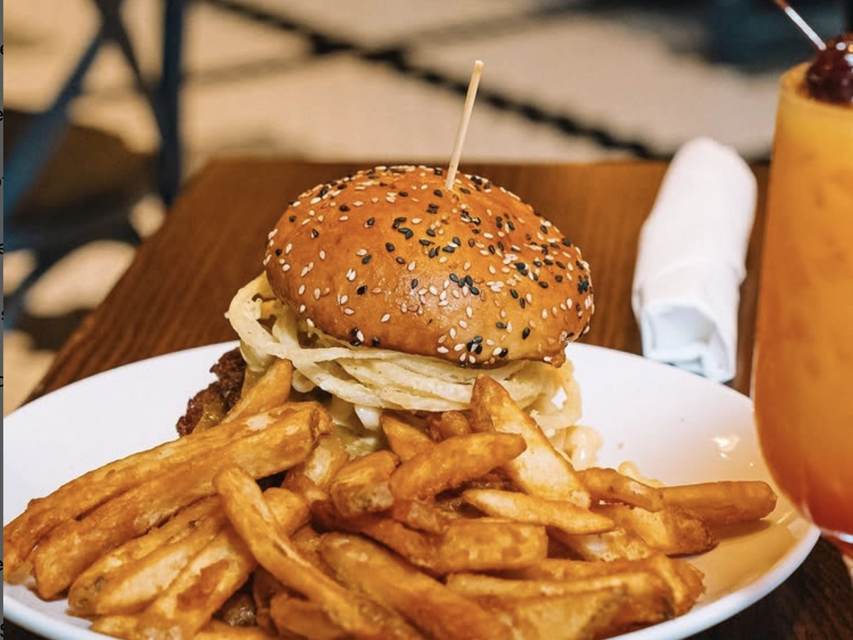 A sesame seed bun burger stacked with crispy onions and melted cheese, served with thick-cut seasoned fries on a white plate, alongside a tropical cocktail garnished with a cherry.