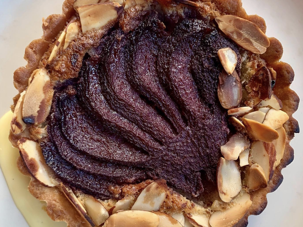 Overhead view of a baked tart topped with sliced poached pear, surrounded by toasted almond slivers and served on a pool of pale custard.