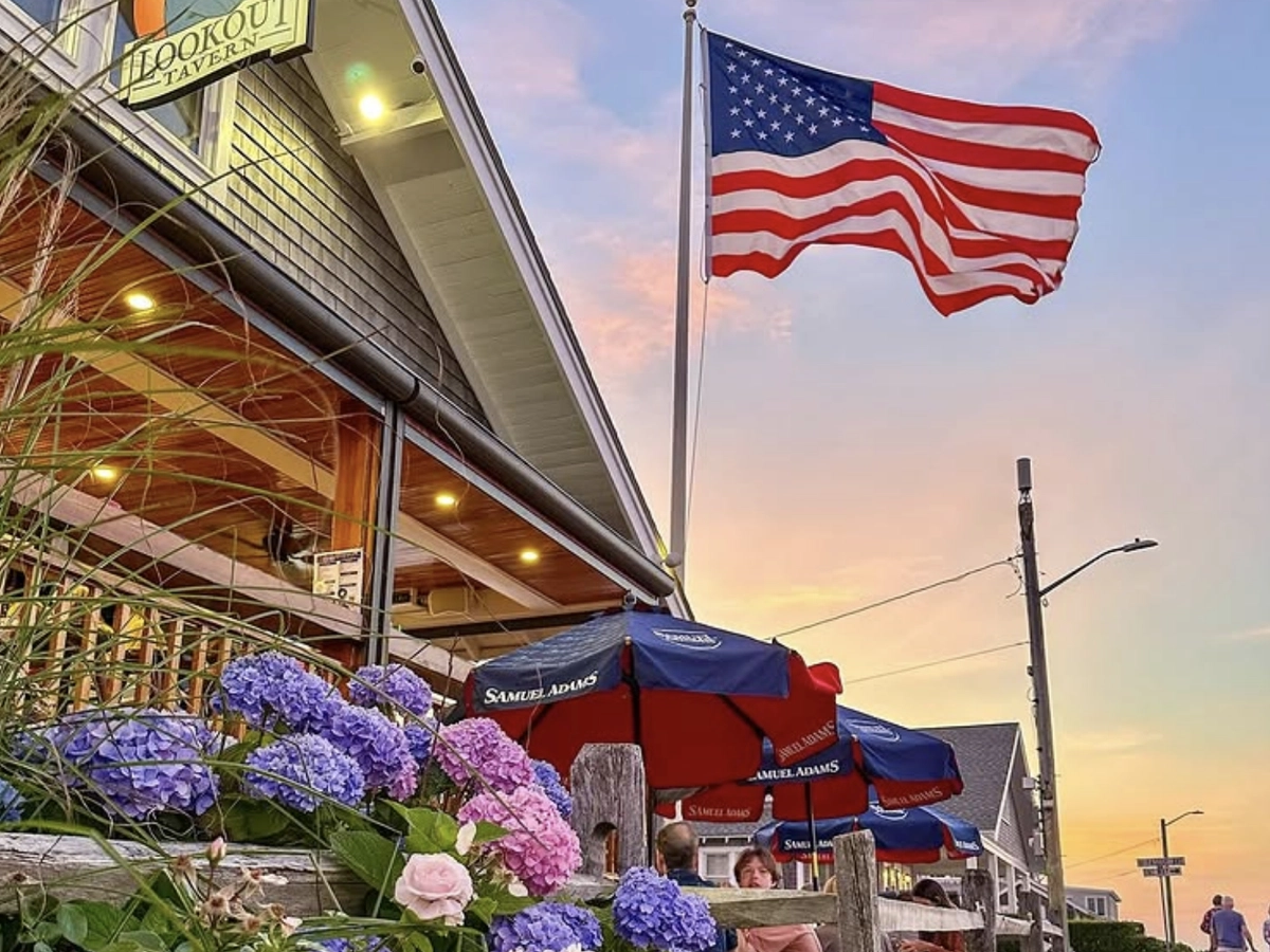 Lookout Tavern at sunset with a large American flag waving above outdoor seating, surrounded by blooming hydrangeas and Samuel Adams patio umbrellas.
