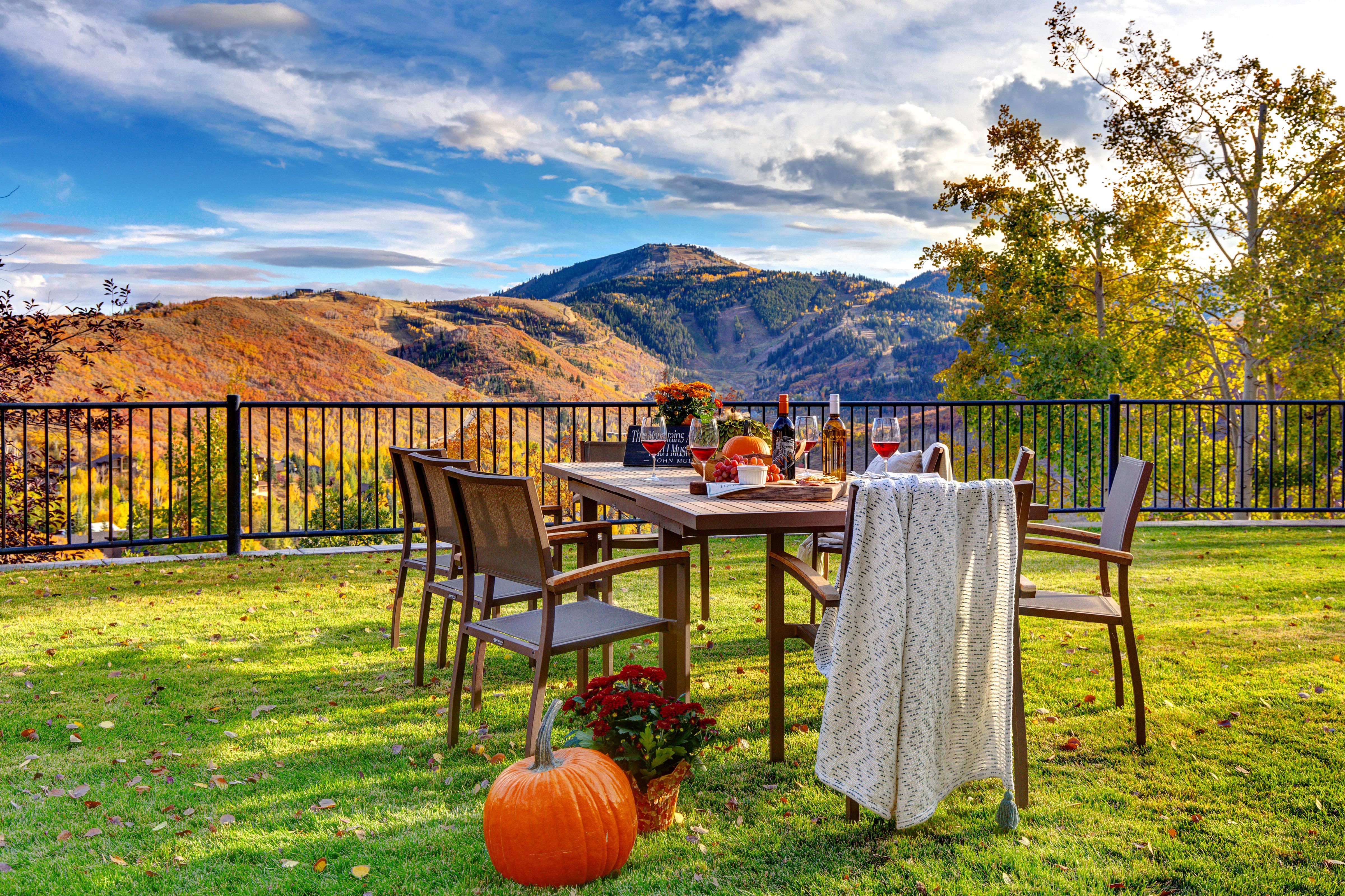 An outdoor dining table set on a grassy lawn with fall-themed decor, wine, and a pumpkin, overlooking a stunning mountain range covered in vibrant autumn foliage under a partly cloudy sky.