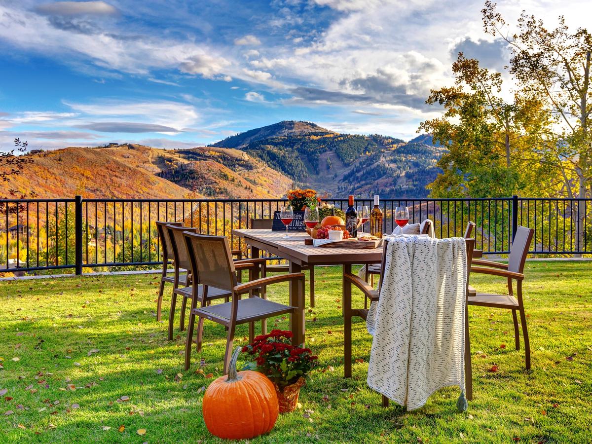 An outdoor dining table set on a grassy lawn with fall-themed decor, wine, and a pumpkin, overlooking a stunning mountain range covered in vibrant autumn foliage under a partly cloudy sky.