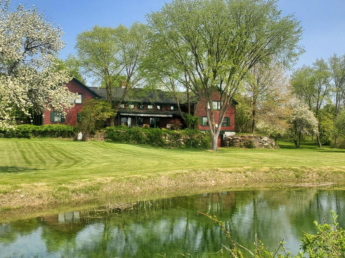 A charming red country home with black trim sits behind a lush green lawn and a reflective pond, surrounded by blooming trees and vibrant spring foliage under a clear blue sky.