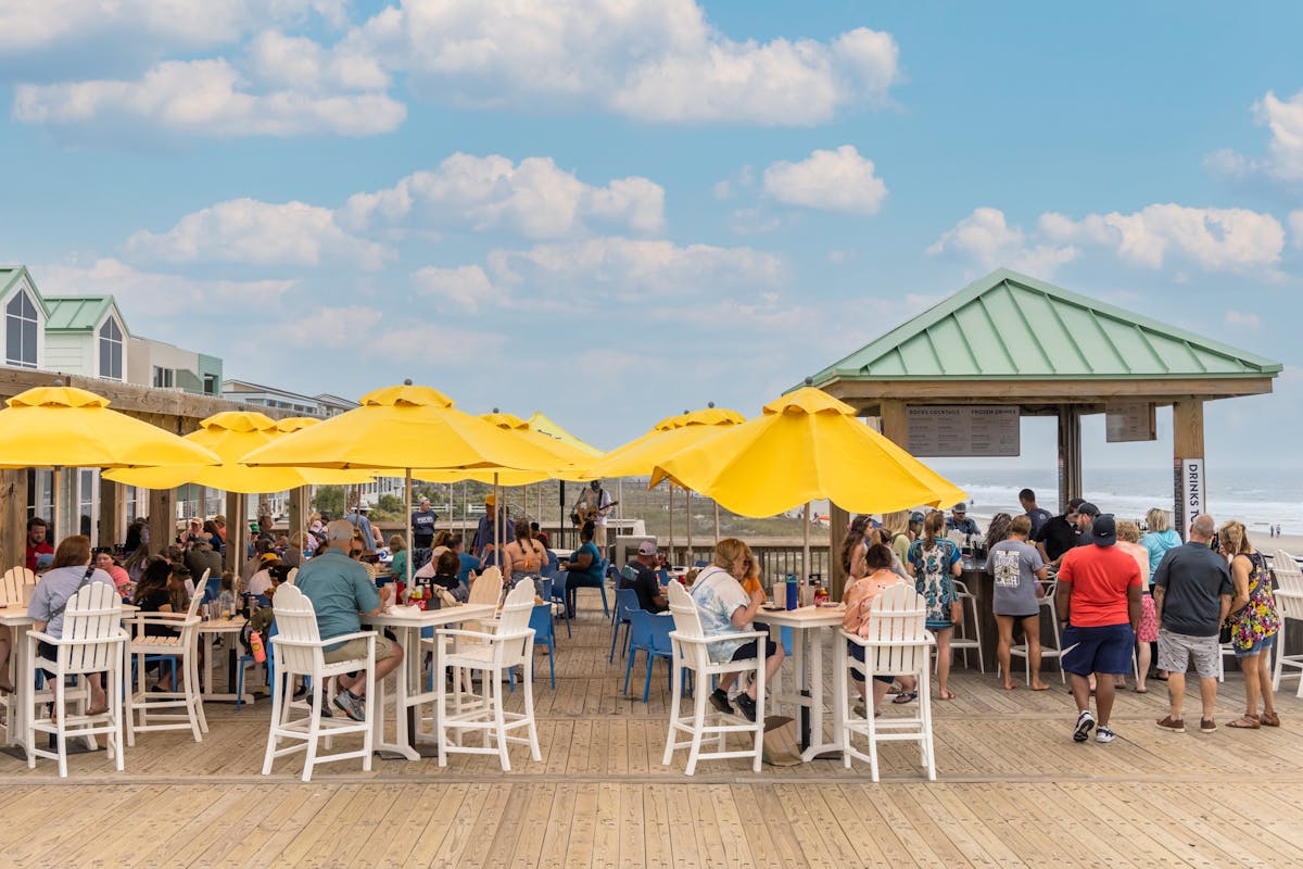 Guests gather around the open-air bar, enjoying drinks with ocean views in the background. The casual seating and beachside setup make it a popular spot for sunset cocktails. It captures the fun, social vibe of dining right on the pier.