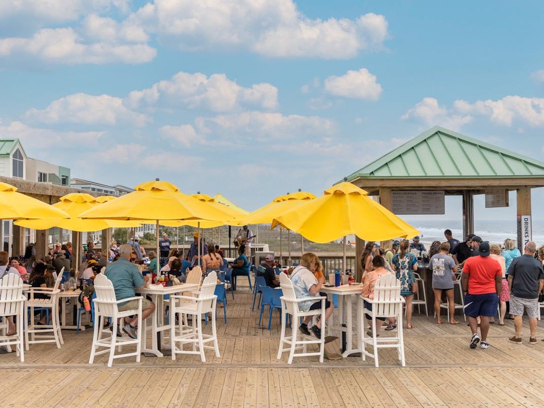 Guests gather around the open-air bar, enjoying drinks with ocean views in the background. The casual seating and beachside setup make it a popular spot for sunset cocktails. It captures the fun, social vibe of dining right on the pier.