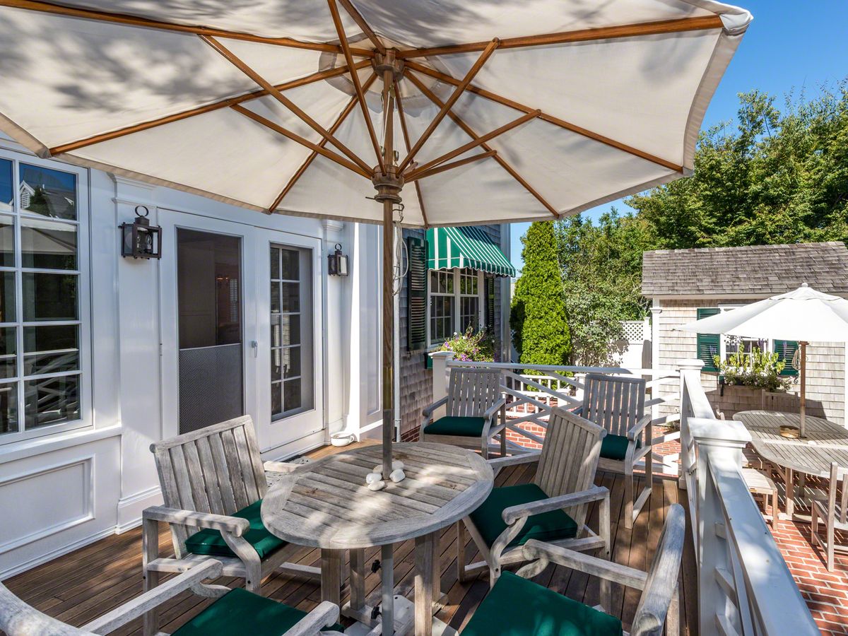 A charming outdoor deck features weathered wood furniture with green cushions, large umbrellas for shade, and overlooks a brick patio and garden area surrounded by greenery.
