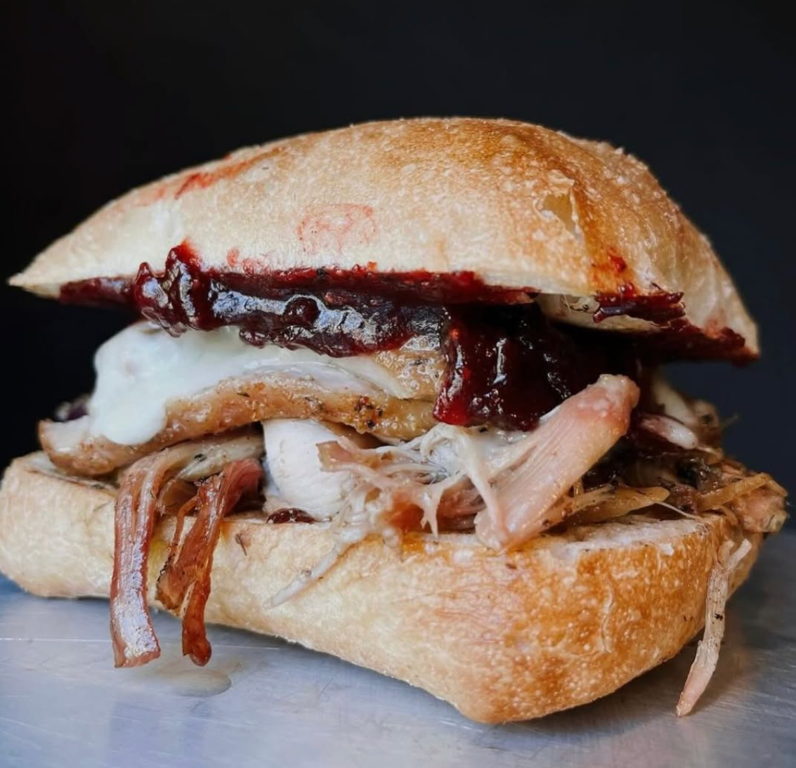A close-up of a rustic pulled-pork sandwich on a crusty ciabatta roll, layered with melted cheese and glossy dark barbecue sauce dripping slightly over the edges.