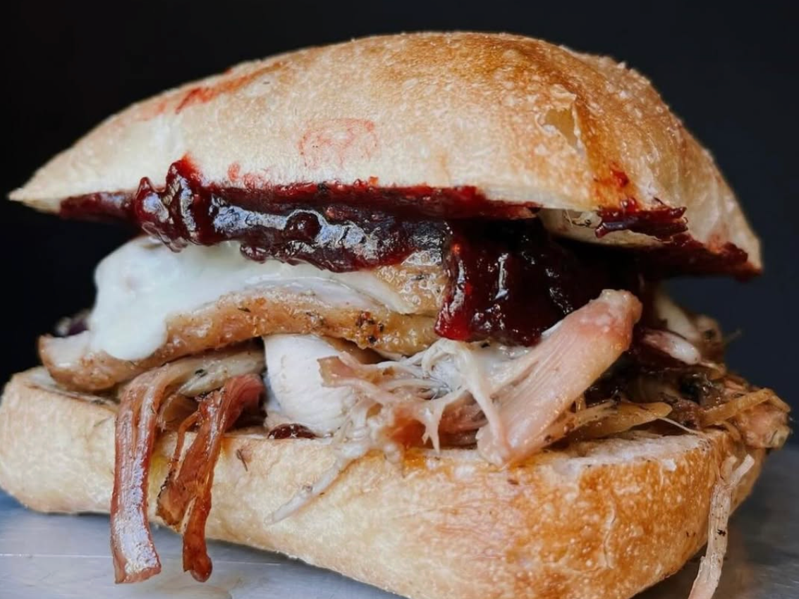 A close-up of a rustic pulled-pork sandwich on a crusty ciabatta roll, layered with melted cheese and glossy dark barbecue sauce dripping slightly over the edges.