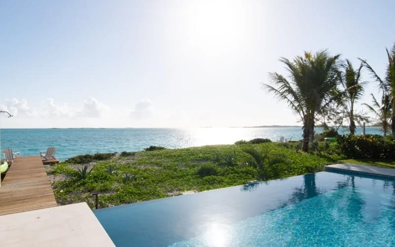 An infinity pool overlooks lush greenery and the sparkling turquoise ocean, with the sun shining bright over the horizon and palm trees swaying nearby.