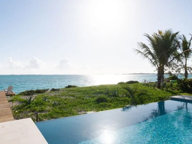 An infinity pool overlooks lush greenery and the sparkling turquoise ocean, with the sun shining bright over the horizon and palm trees swaying nearby.