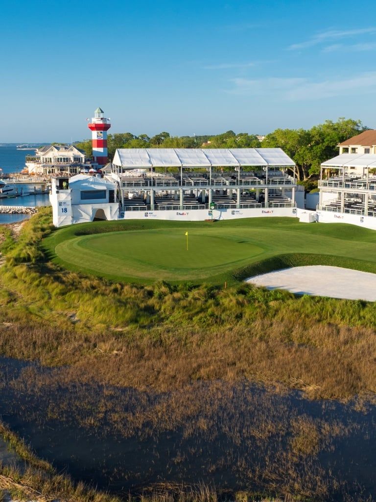 The iconic 18th hole at Harbour Town Golf Links on Hilton Head Island features a pristine green and sand bunker steps from the famous red-and-white striped lighthouse, with tournament grandstands and marina boats visible along the Calibogue Sound.
