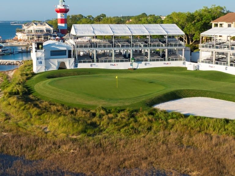 The iconic 18th hole at Harbour Town Golf Links on Hilton Head Island features a pristine green and sand bunker steps from the famous red-and-white striped lighthouse, with tournament grandstands and marina boats visible along the Calibogue Sound.