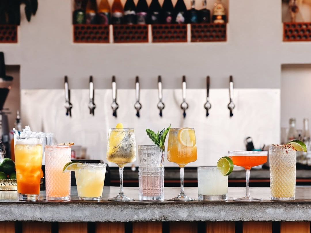 A colorful lineup of cocktails sits on the bar at Burial Beer Co. in Asheville, with sleek taps and bottle displays in the background. Known for its bold beers and creative vibe, this brewery also shines with its craft cocktails and stylish setting.