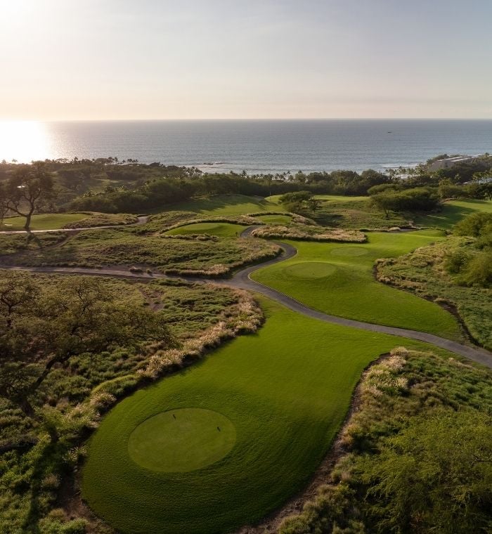 An aerial view of Mauna Kea Golf Course on the Big Island of Hawaii reveals sweeping holes cut through native lava fields and golden grasses, with the Pacific Ocean glinting in the background at sunse