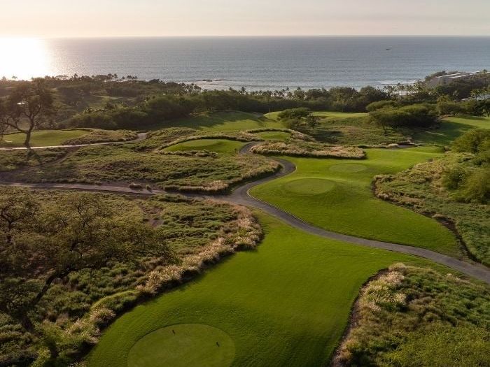 An aerial view of Mauna Kea Golf Course on the Big Island of Hawaii reveals sweeping holes cut through native lava fields and golden grasses, with the Pacific Ocean glinting in the background at sunse