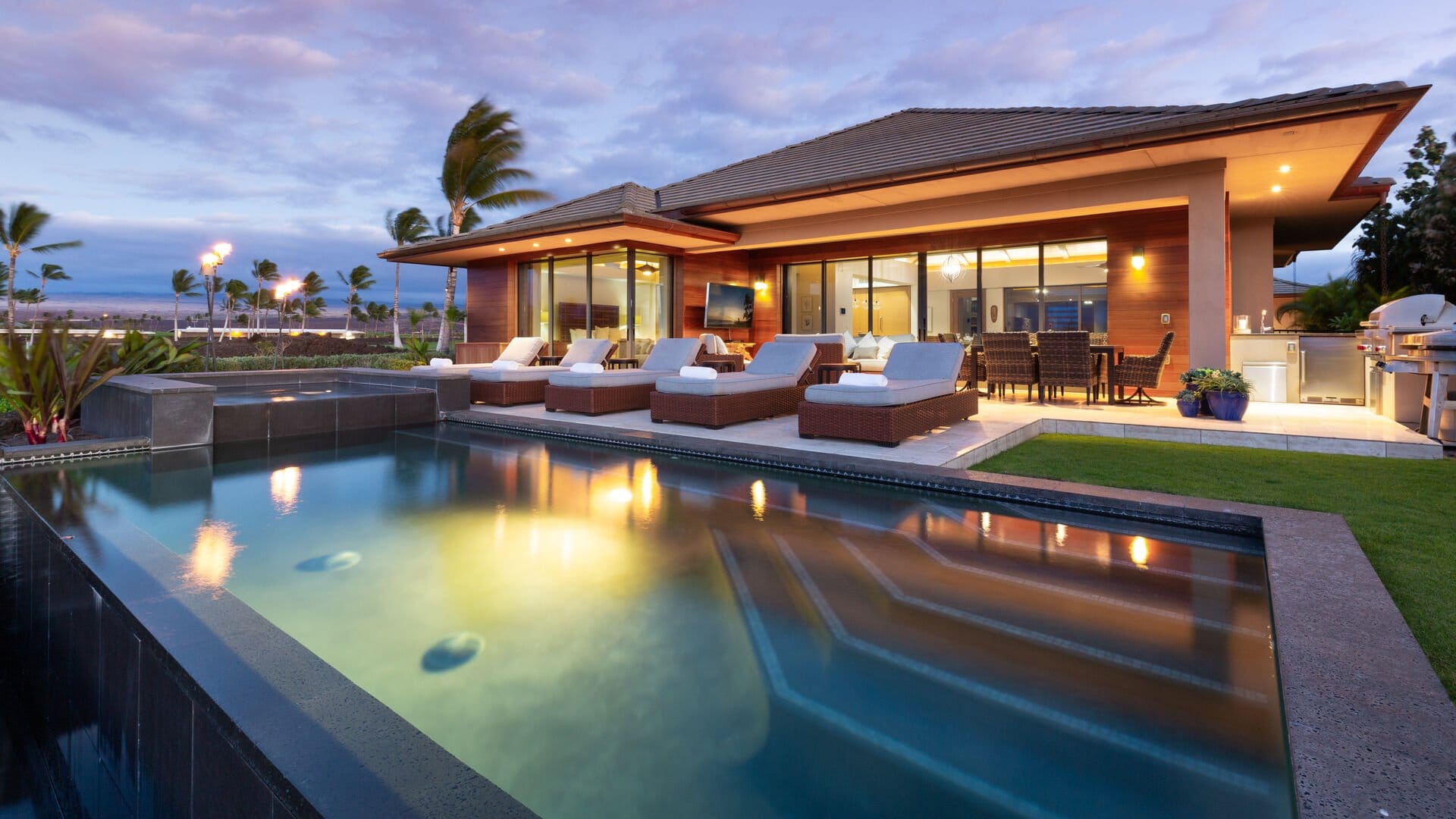 The stunning outdoor living area of Elegant Escape on the Big Island of Hawaii features a sleek lap pool with a spillover spa reflecting the warm glow of the home at twilight, flanked by cushioned sun loungers and an outdoor dining set. Floor-to-ceiling glass walls open the contemporary home to swaying palms and a dramatic purple Hawaiian sunset sky.