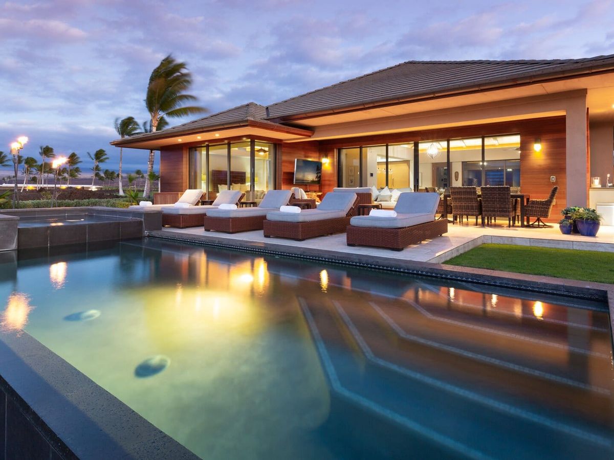 The stunning outdoor living area of Elegant Escape on the Big Island of Hawaii features a sleek lap pool with a spillover spa reflecting the warm glow of the home at twilight, flanked by cushioned sun loungers and an outdoor dining set. Floor-to-ceiling glass walls open the contemporary home to swaying palms and a dramatic purple Hawaiian sunset sky.