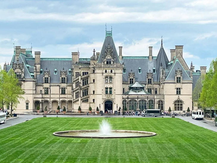 A grand château-style mansion with a central fountain and expansive green lawn in front, framed by trees and a cloudy sky.