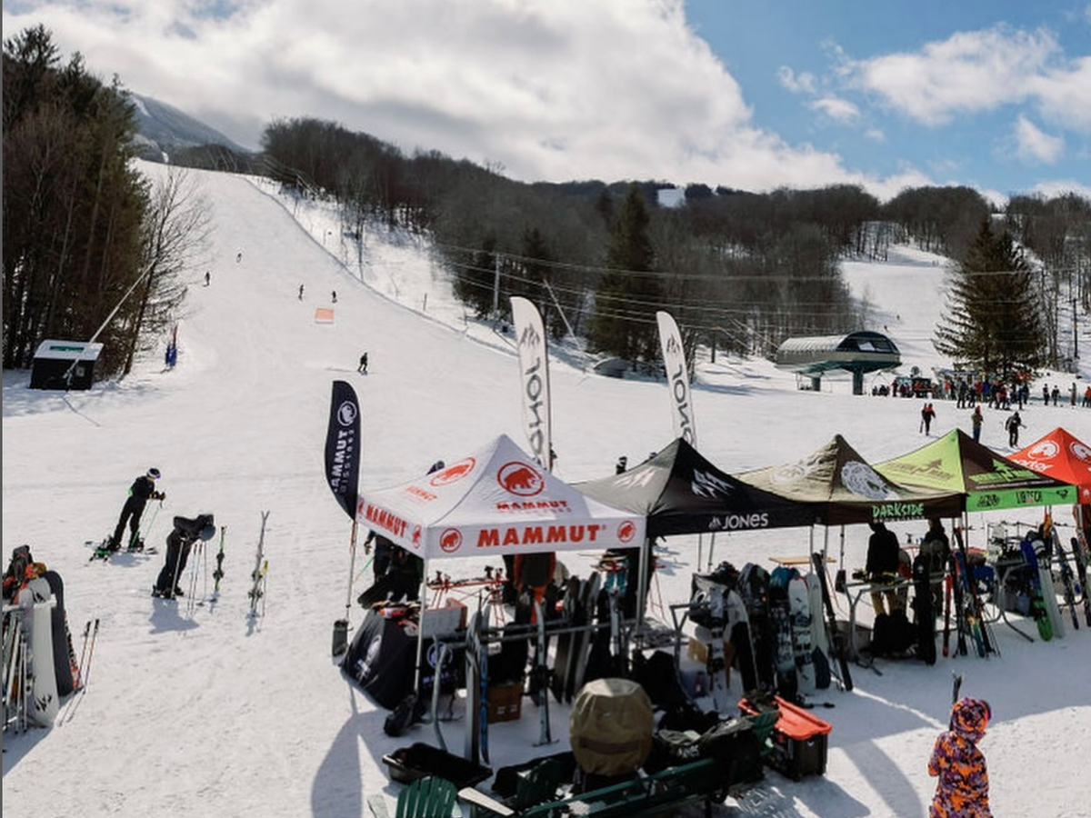 A lively ski resort base features vendor tents, skiers preparing gear, and a snowy slope in the background under a bright blue sky.