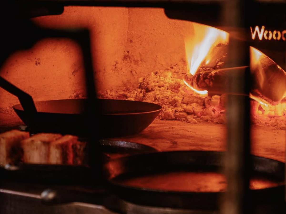 Thick slices of bread toast in cast iron pans inside a wood-fired oven, with flames and glowing embers providing a rustic, fiery backdrop.
