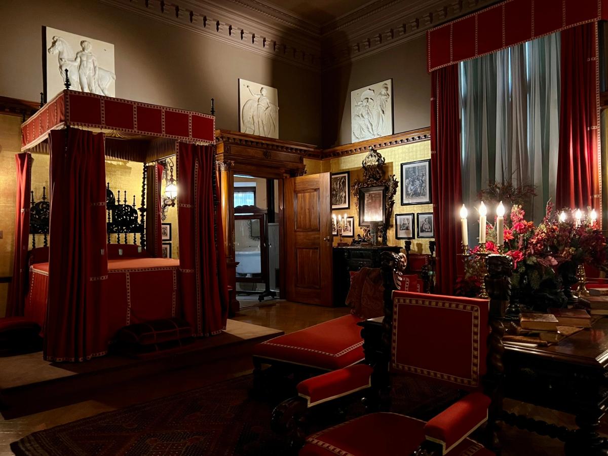 Lavishly decorated historic bedroom featuring a grand red canopy bed, ornate wooden furniture, candlelit candelabras, and richly adorned walls with classical art and deep red drapes.