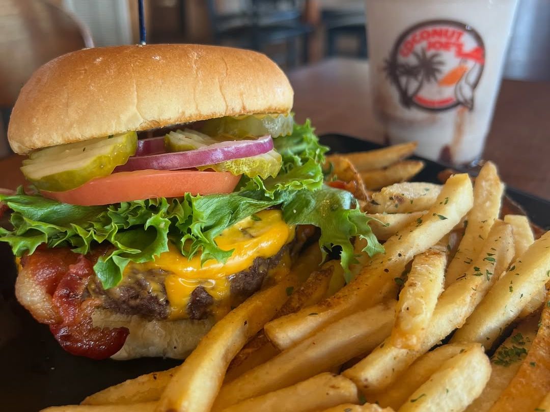 Juicy cheeseburger stacked high with toppings, crispy fries, and a frozen drink in the background at Coconut Joe’s Beach Grill. This is comfort food done beach-style, and it hits the spot every time.