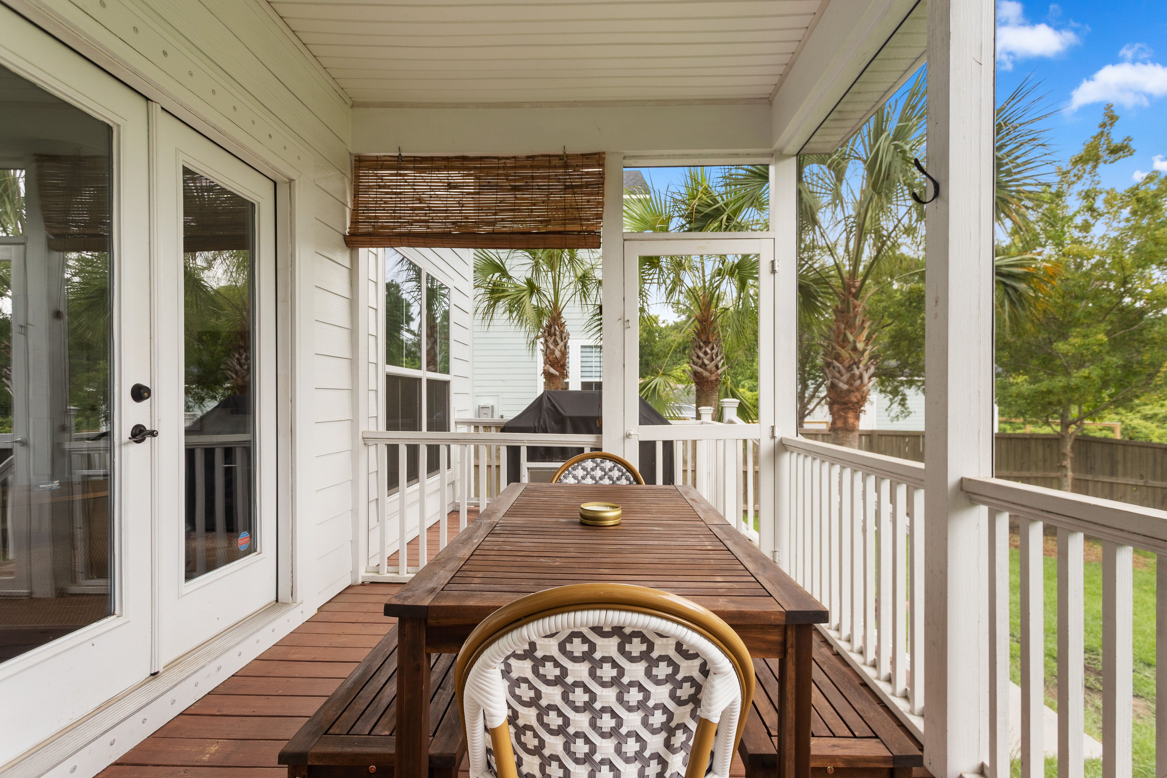 A cozy screened-in porch features a wooden dining table with patterned chairs, surrounded by white railings and palm trees in the backyard.