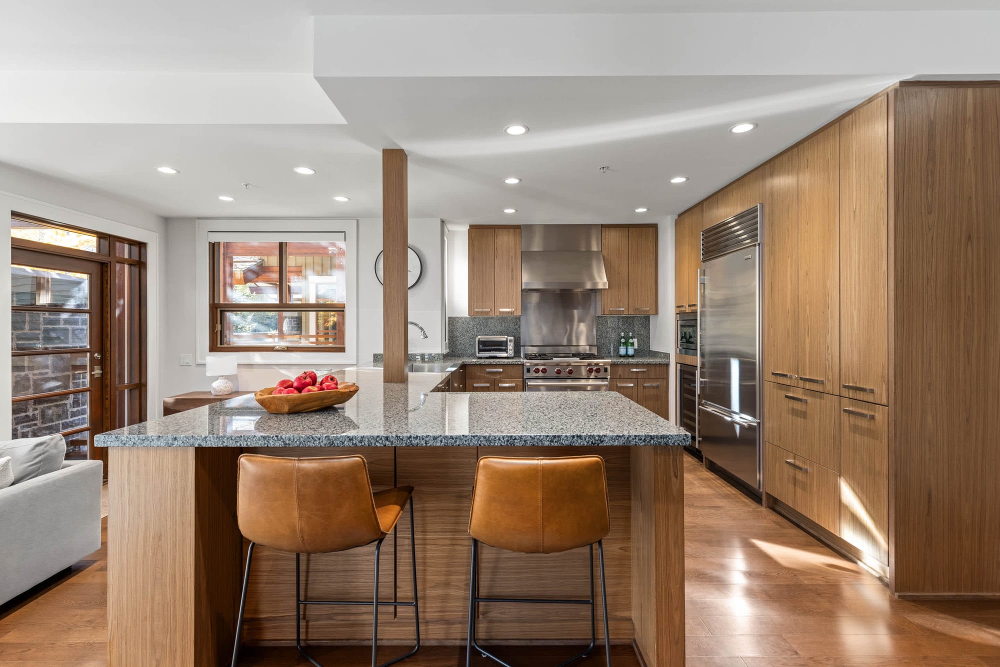 Warm, contemporary kitchen with wood cabinetry, stainless steel appliances, a granite island with leather barstools, and bright recessed lighting throughout.