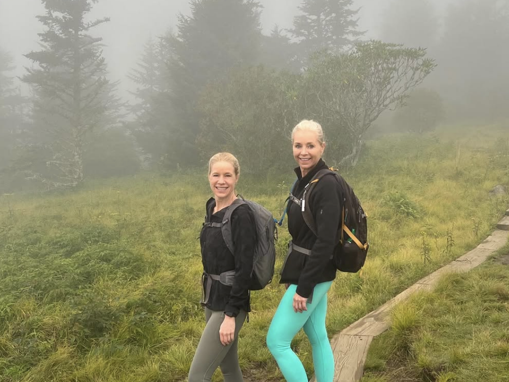 Two women in hiking gear smile on a foggy mountain trail surrounded by misty evergreens and grassy terrain in the Great Smoky Mountains.